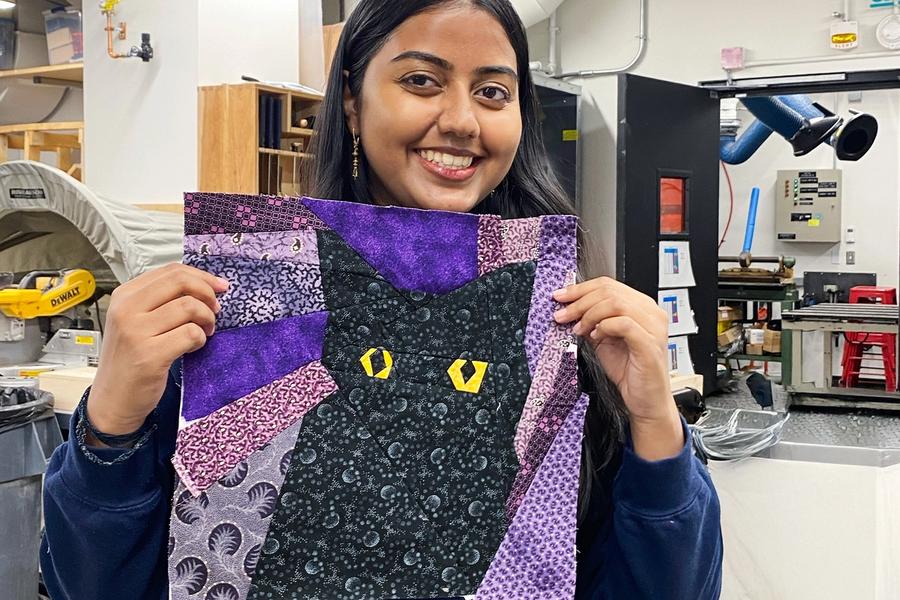 Person holding up a finished quilt block featuring a black cat with bright yellow eyes, surrounded by purple, lavender, and patterned fabric shapes. The background appears to be a makerspace workshop.